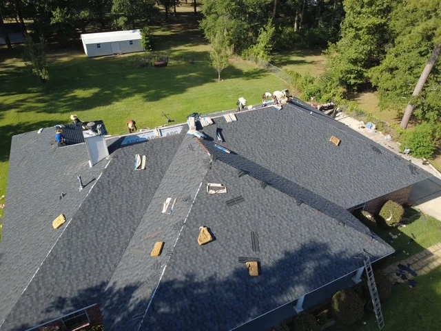 Aerial view of new charcoal gray architectural shingles installed on multi-section residential roof with workers and equipment visible